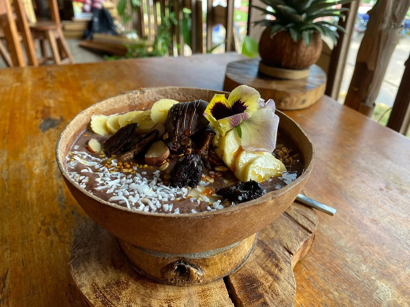 A wooden bowl containing a chocolate dessert topped with sliced bananas, edible flowers, nuts, and coconut flakes, set on a wooden table with a rustic background.