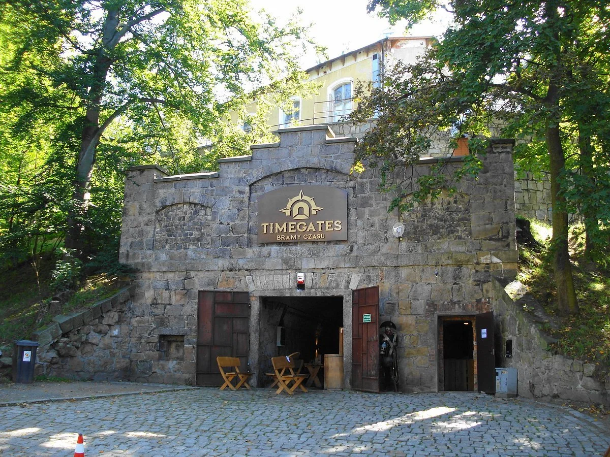A stone building with a sign that reads "Timegates Bramy Czasu" above an arched entrance. The entrance has wooden doors, with some outdoor furniture and bicycles visible. Trees surround the building, with sunlight filtering through the leaves.
