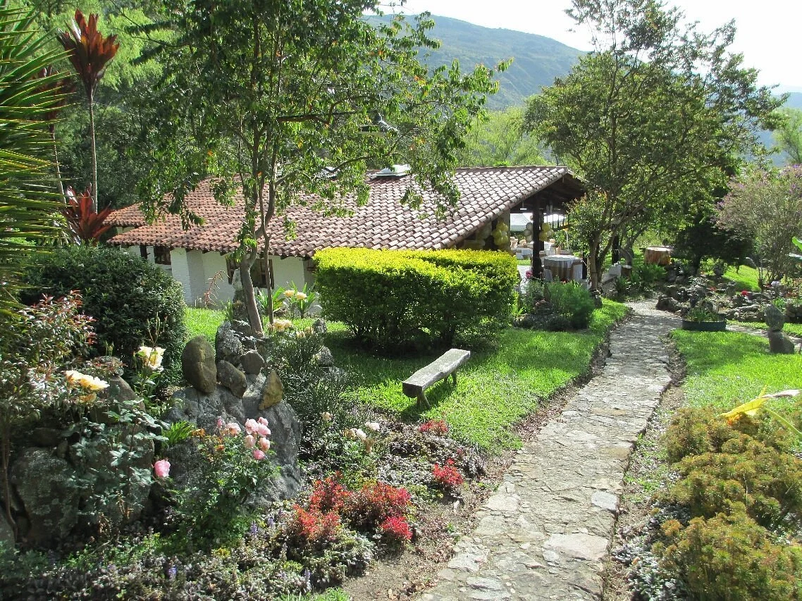 A scenic garden with a stone pathway, various green trees, bushes, and colorful flowers, with a house featuring a tiled roof in the background and mountains in the distance.