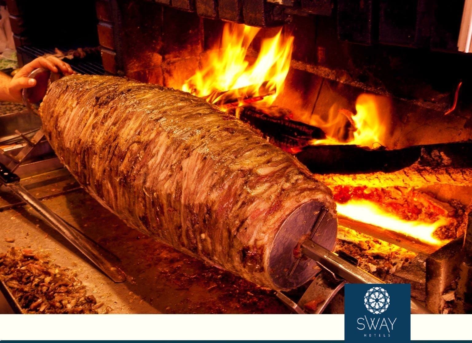 A large roasted meat on a rotisserie spit in front of an open fire in a brick oven, with a person adjusting it.