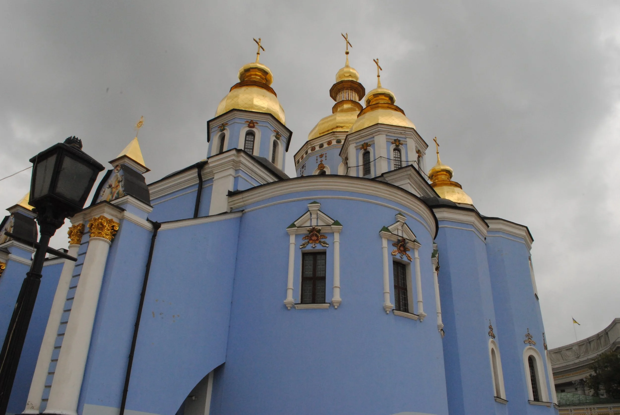 A church with a blue exterior and golden domes topped with crosses, under a cloudy sky, with a street lamp in the foreground.