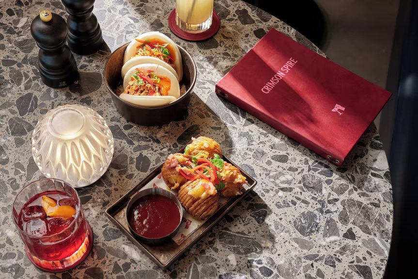 A table with three tacos topped with vegetables, a small dish of red sauce, and a drink with ice and a lemon slice. There are also a pepper grinder, a salt shaker, a decorative white vase, a glass of yellow beverage, and a red menu titled 'Crimson Sp