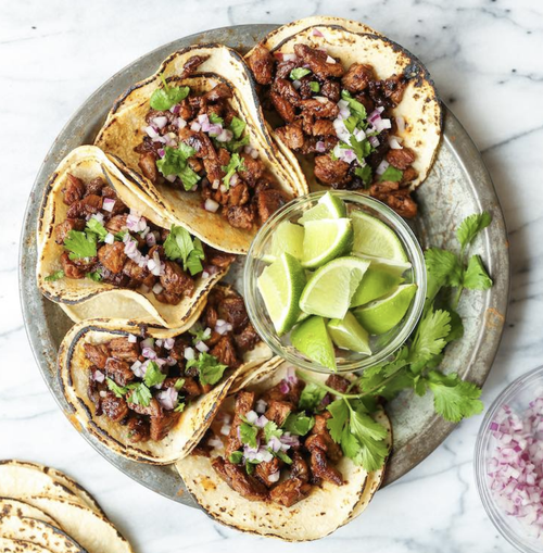 Four beef tacos garnished with chopped onions and cilantro, with a bowl of lime wedges on a silver platter.