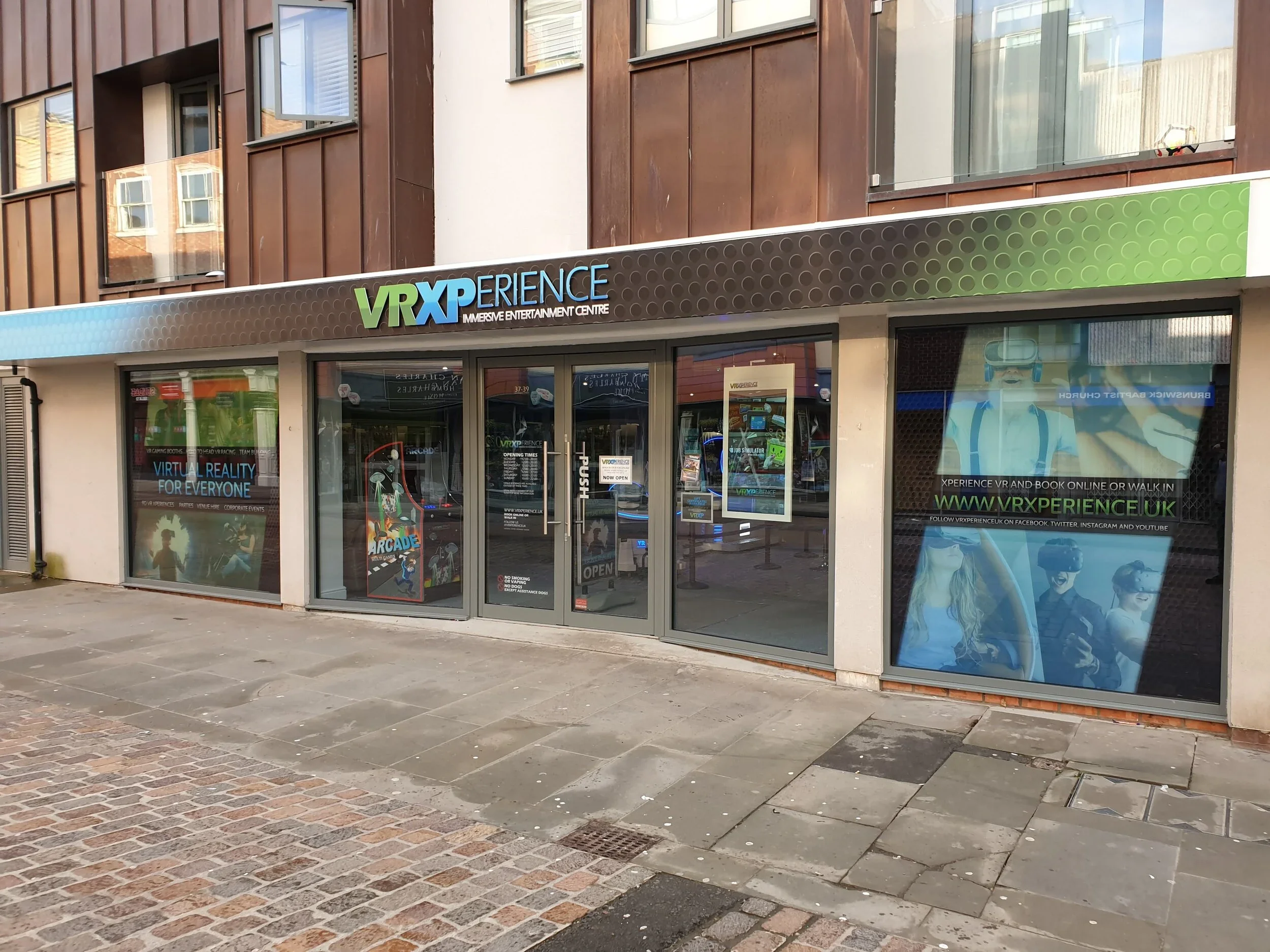 The storefront of VRXperience, an immersive entertainment center, featuring large windows with promotional posters and a colorful sign above the entrance. The building has a brown and beige exterior, with a glass door and window displays.