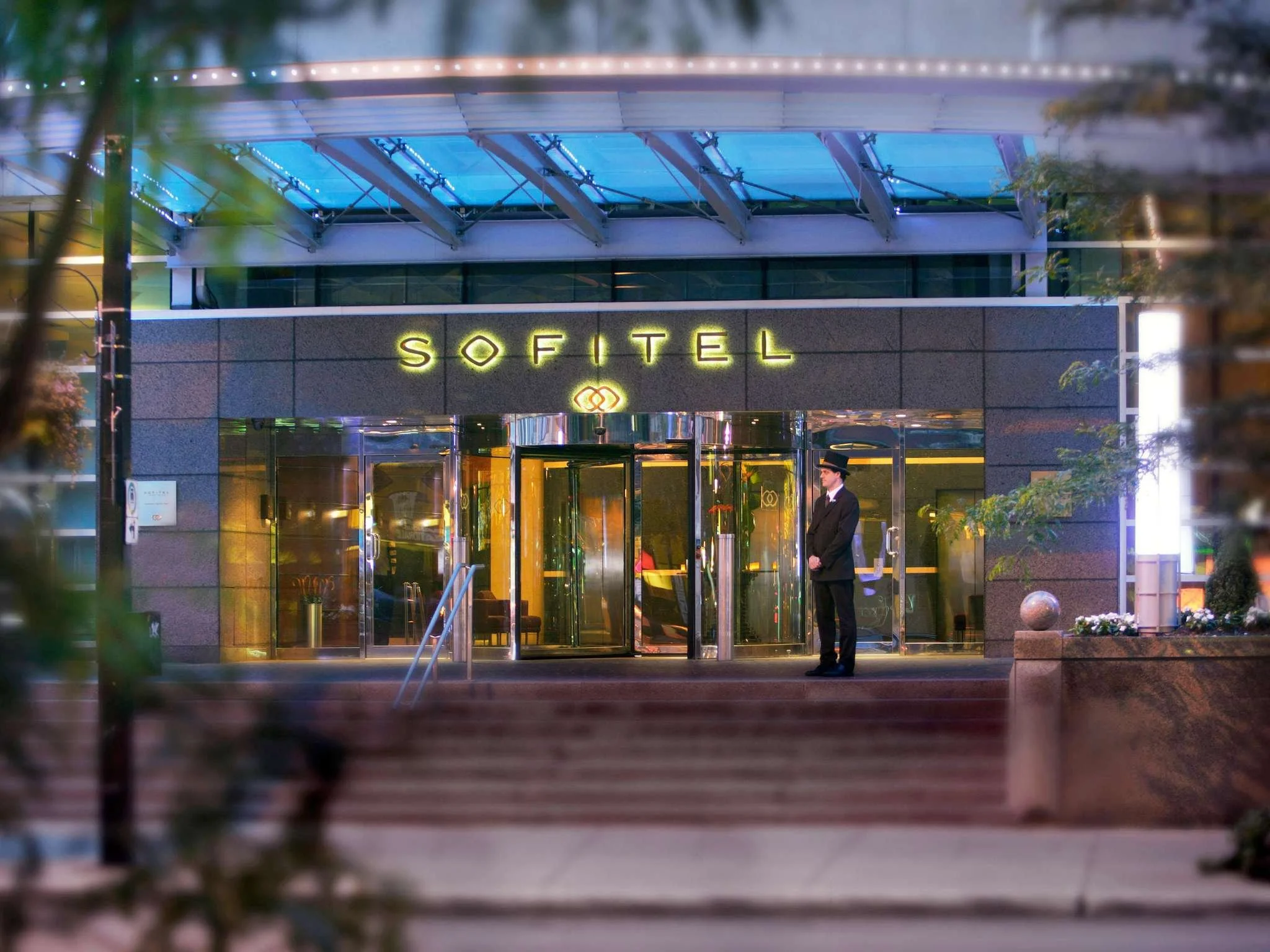Entrance to Sofitel hotel with a doorman standing guard, illuminated sign reading 'SOFITEL', glass doors, steps leading up to the entrance, and trees partially visible on the sides.