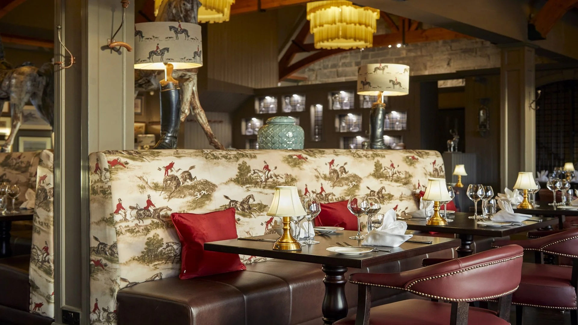 Elegant restaurant dining area with decorated booth seating featuring a countryside riding scene, set with white napkins, wine glasses, and gold lamps on dark wood tables. Background includes warm lighting, wood accents, and display cabinets.