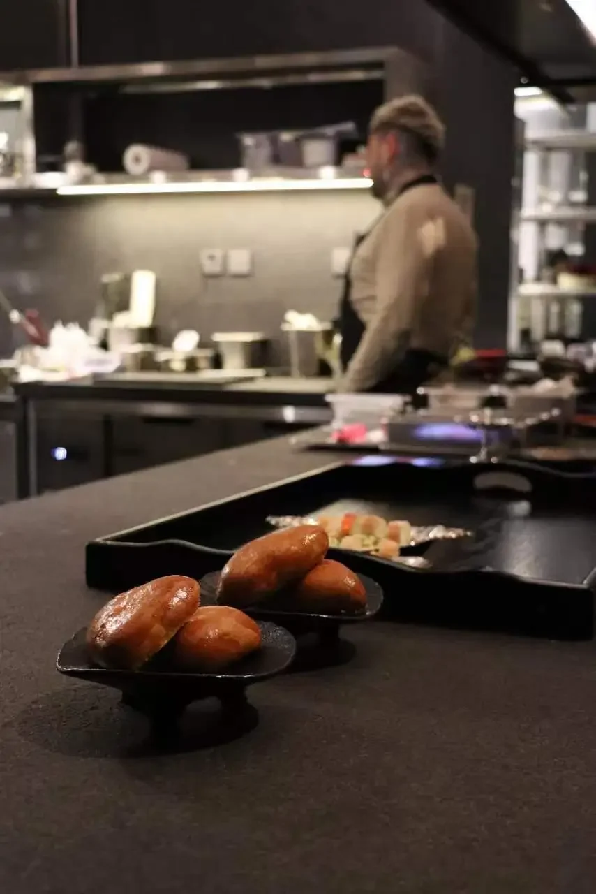 Close-up of three glazed donuts on black stands in a restaurant setting, with a blurred chef working in the background.