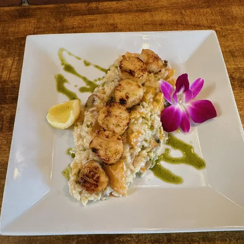 Seafood dish with scallops, rice, and vegetables, garnished with lemon wedge, purple orchid, and drizzled green sauce on a white square plate