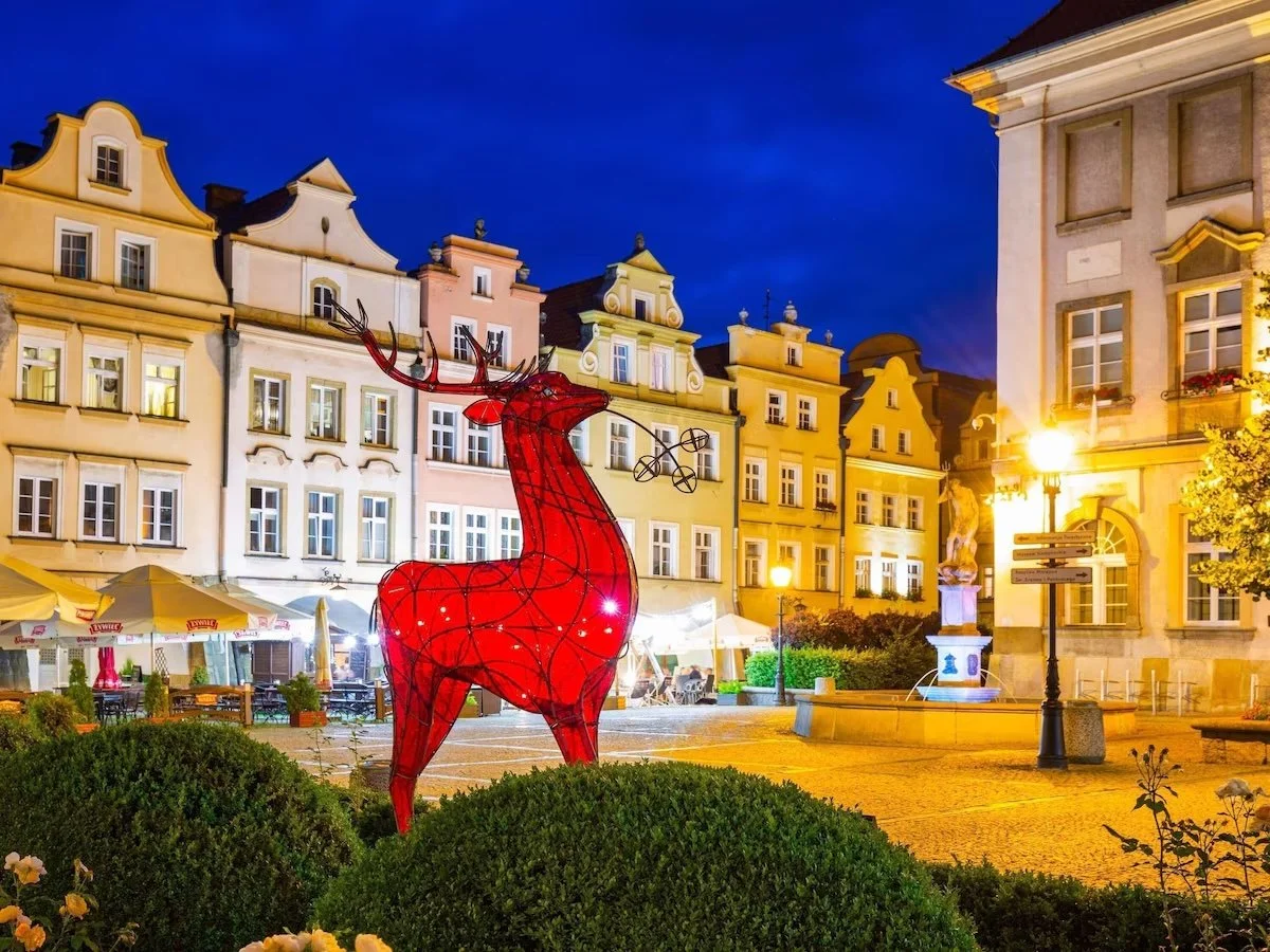 A large illuminated red reindeer sculpture in a European city square at night, with colorful historic buildings, streetlights, and outdoor seating areas in the background.