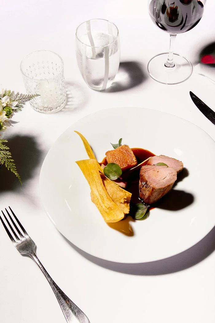 Elegant dinner plate with a portion of cooked meat, garnished with greens, served with a slice of yellow vegetable and a sauce, set on a white table with wine, water, and glassware.