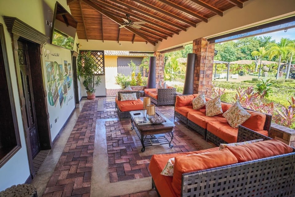 Covered outdoor seating area with wicker couches and orange cushions, brick columns, a brick-paved floor, a wall-mounted TV, and plants, overlooking a lush garden.