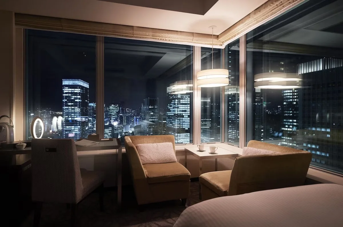 A hotel room with a large window view of a city skyline at night, featuring illuminated skyscrapers, and a seating area with two chairs, a small table, and hanging ceiling lights.
