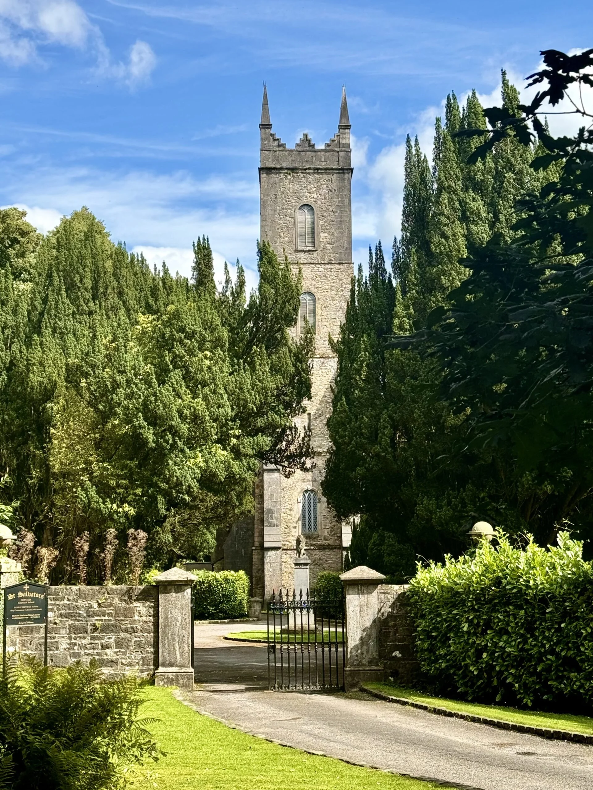 A historic stone church tower with gothic architecture, surrounded by lush green trees and a well-maintained garden, with a black iron gate at the entrance, under a bright blue sky with some clouds.