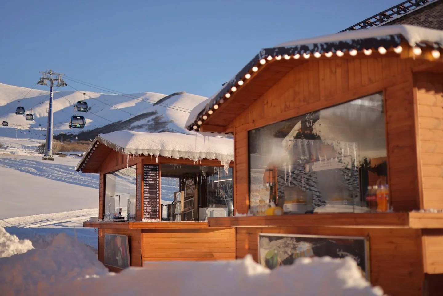Snow-covered wooden ski lodge with a glass window, snow on its roof, in a snowy mountain landscape with ski lifts and ski slopes in the background during daytime.