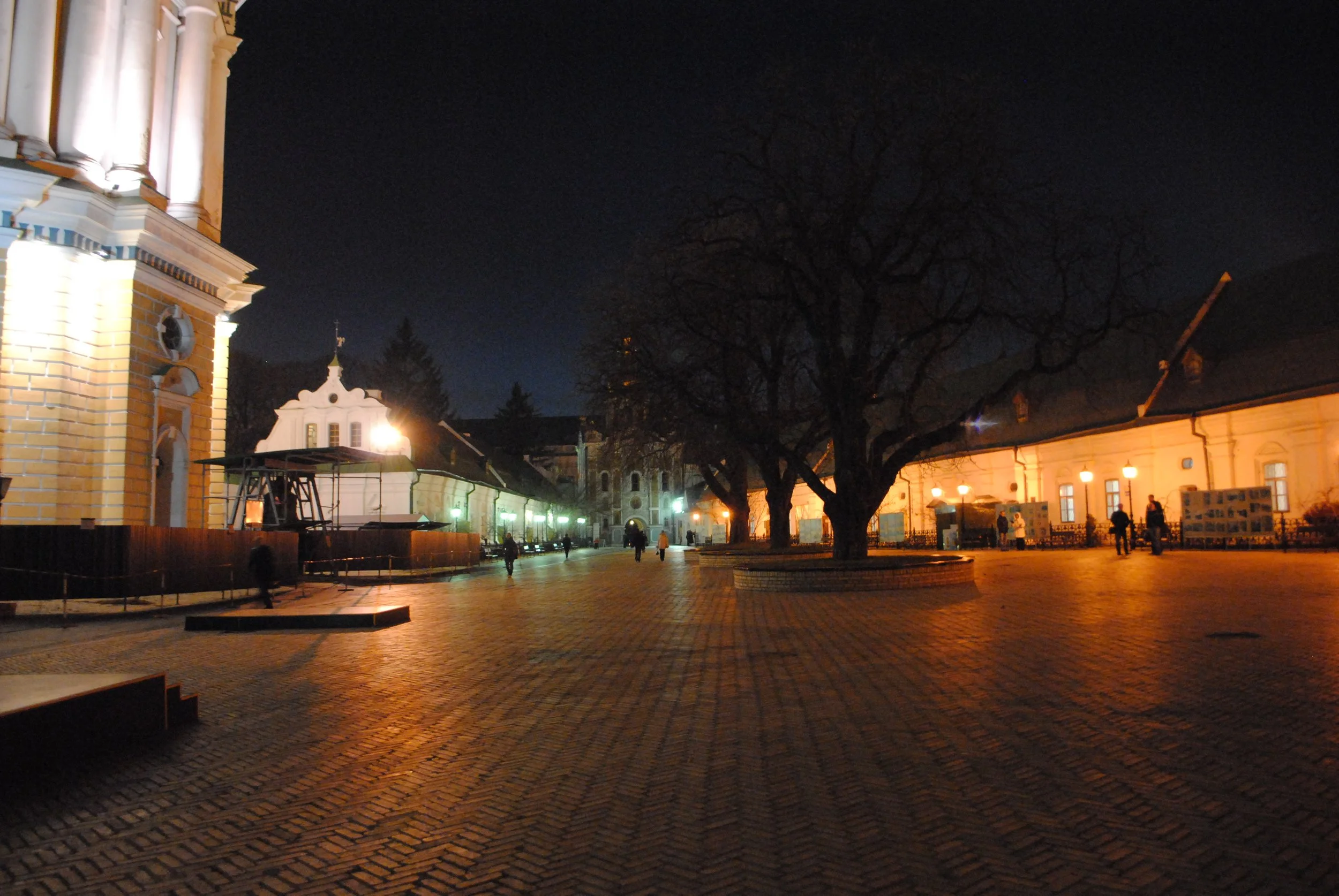 Night scene of a quiet, illuminated European-style town square with cobblestone pavement, historic buildings, leafless trees, and a few people walking.