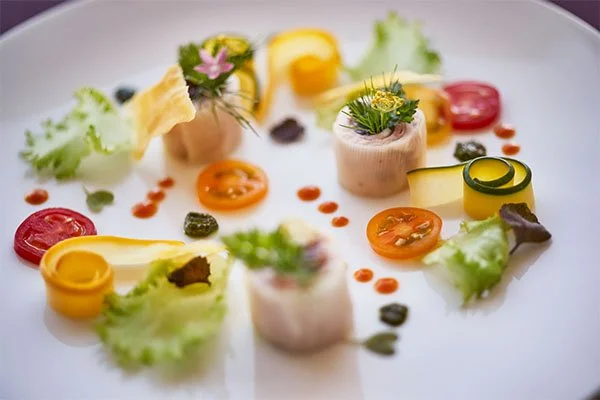 An artistic plate with rolled slices of fish, cherry tomatoes, lettuce, and decorative edible flowers and sauces.