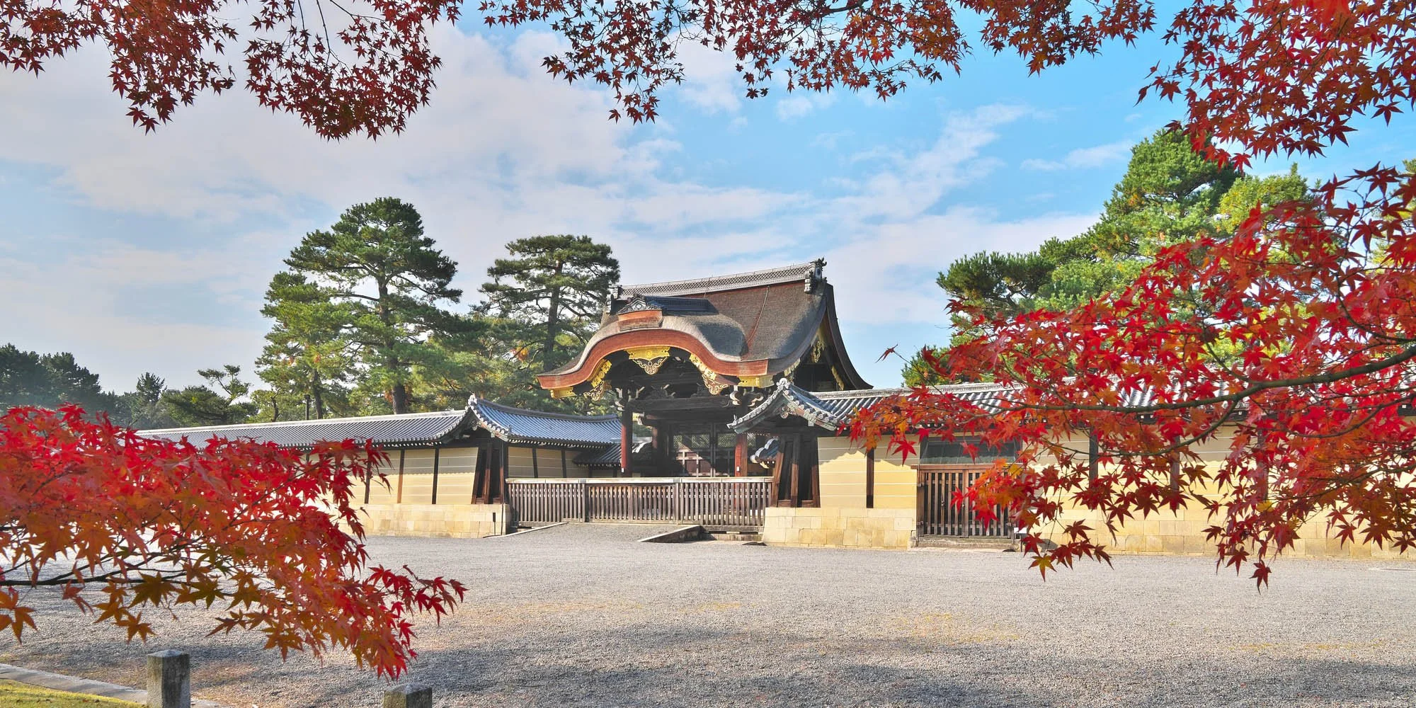 A traditional Japanese temple or shrine surrounded by vibrant red maple leaves and tall green pine trees under a blue sky with scattered clouds.