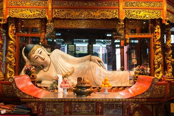 Reclining Buddha statue within a richly decorated temple altar, adorned with offerings and floral arrangements.