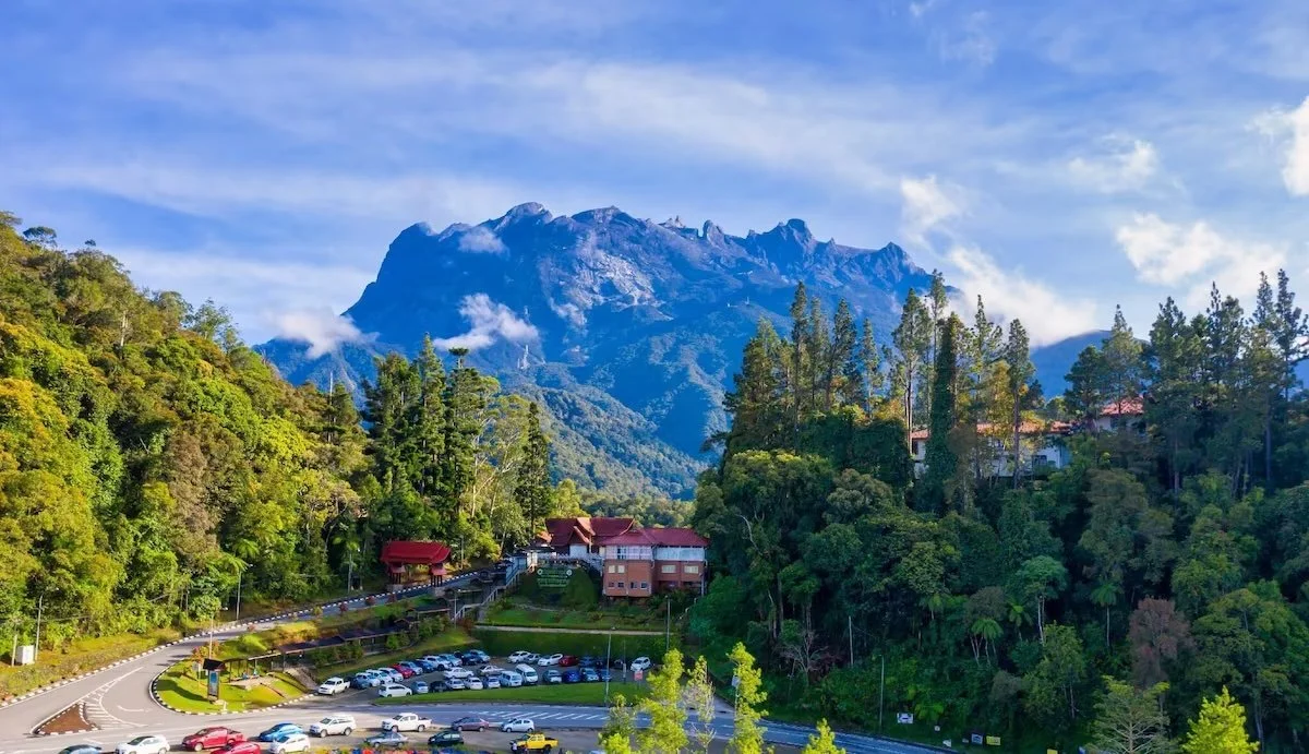Mountain with lush green trees in foreground, buildings on hillside, and a parking lot at the bottom, under a partly cloudy sky.