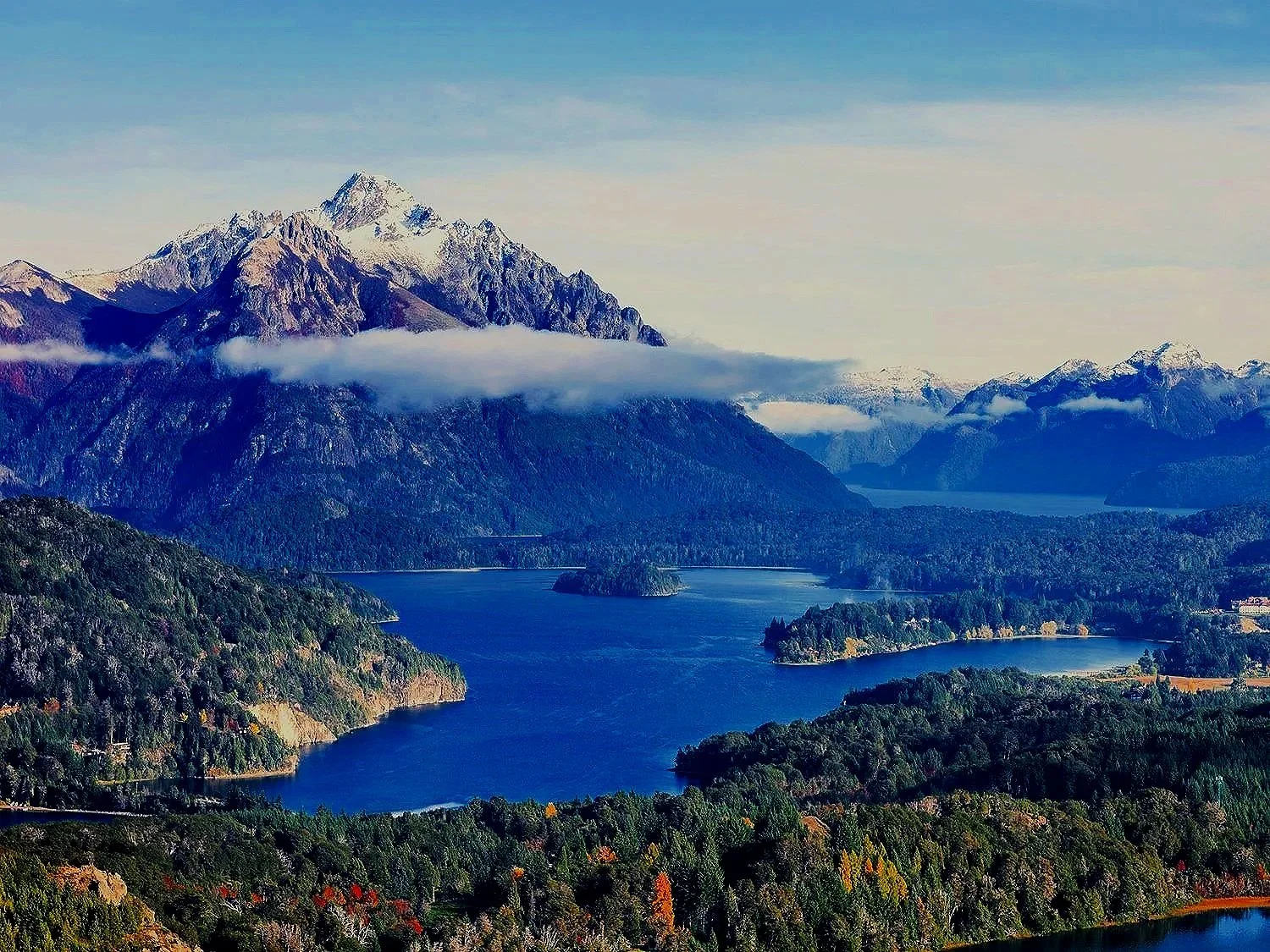 Scenic view of snow-capped mountains over a large lake surrounded by lush green forests and trees.