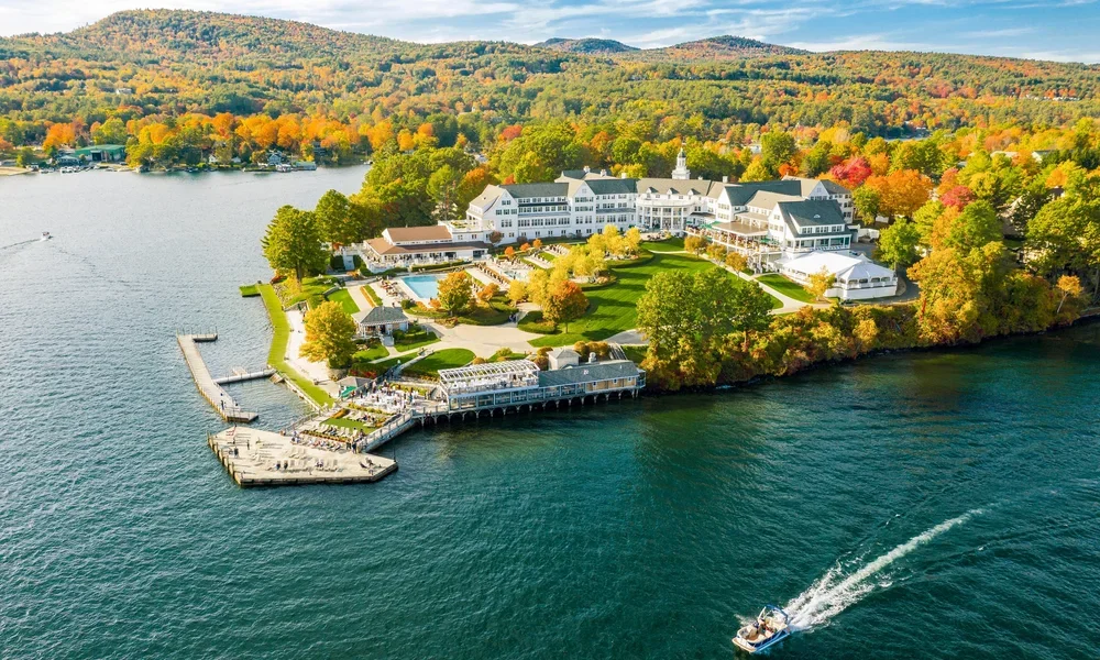 A large lakeside resort with a white main building, outdoor swimming pool, and waterfront pier, surrounded by colorful autumn trees and rolling hills.