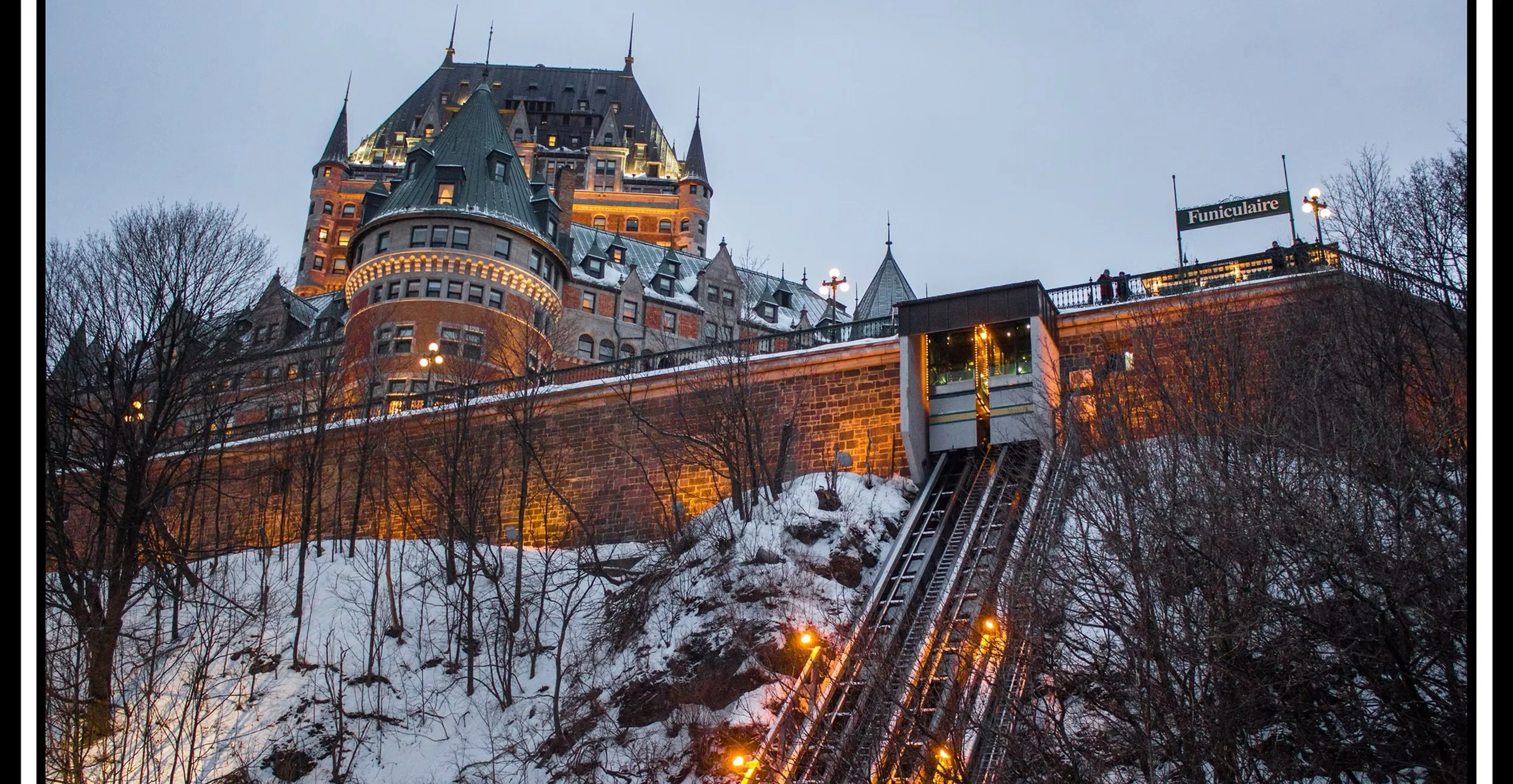 A snow-covered hillside with leafless trees leading up to a castle-like building illuminated at dusk. The building has turrets, towers, and a steeply pitched roof, resembling a fairy tale castle. An elevator shaft with lights is seen descending the h