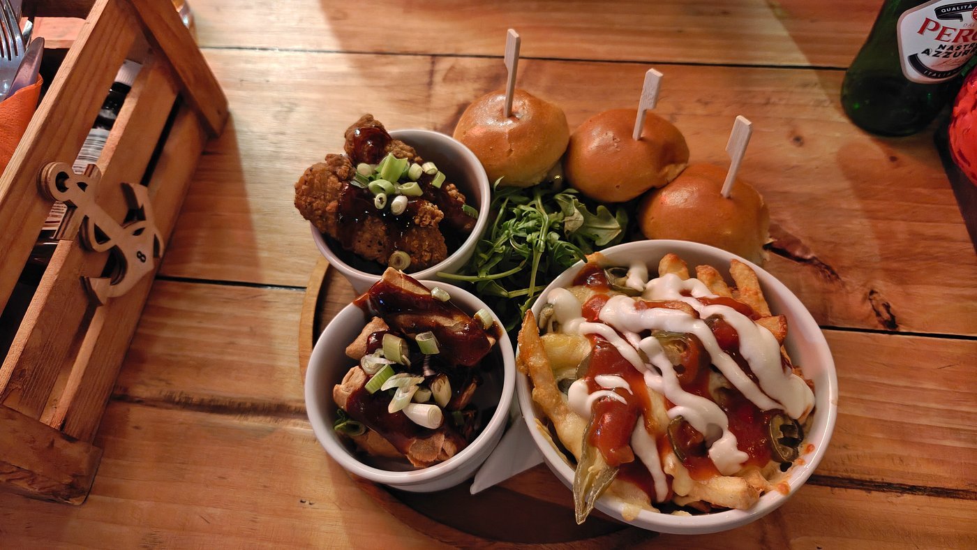 A wooden table with a basket of utensils, a plate of three small sandwiches with sticks, a small bowl of fried chicken with sauce and green onions, a small bowl of beef with green onions, and a bowl of cheesy fries topped with ketchup, ranch, and jal