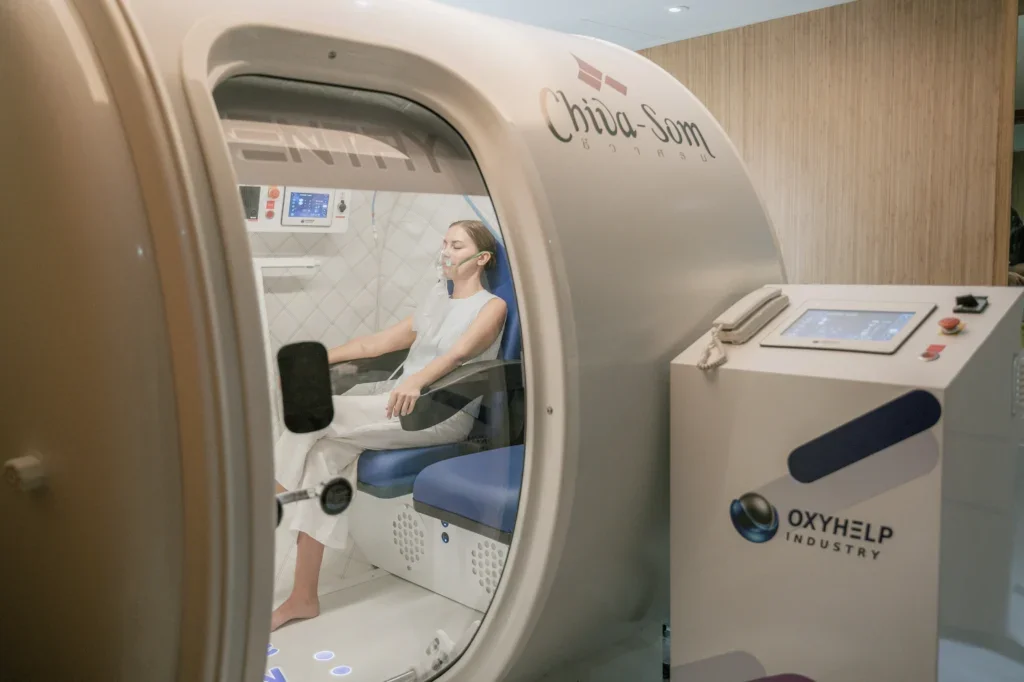 A woman in a hospital gown undergoing oxygen therapy inside a hyperbaric oxygen chamber with the label 'Chiba-Som' on the chamber and 'OXYHELP INDUSTRY' on a nearby control unit.