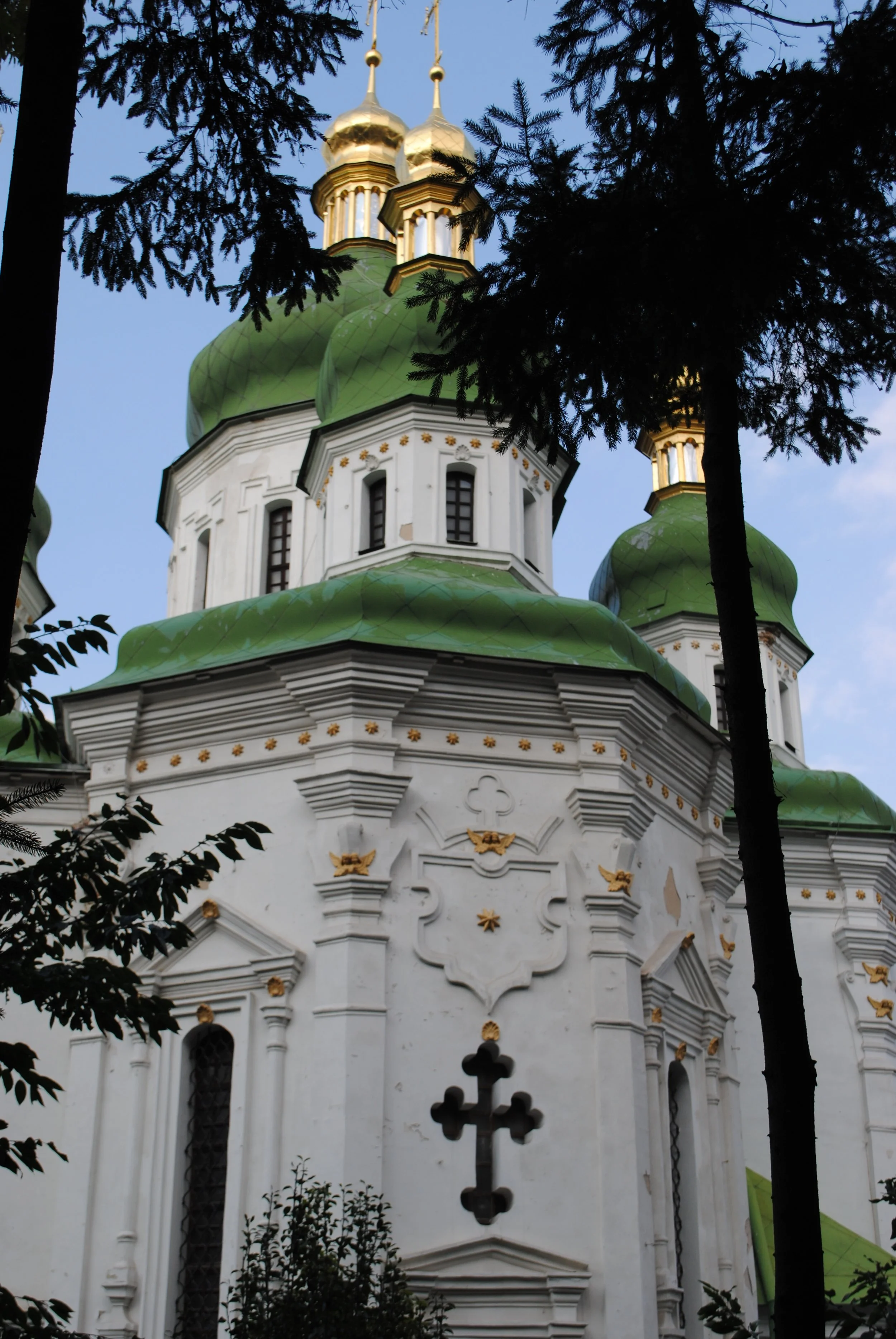 View of a church or cathedral with white ornate walls, gold accents, and green domes topped with gold crosses, surrounded by dark trees against a blue sky.