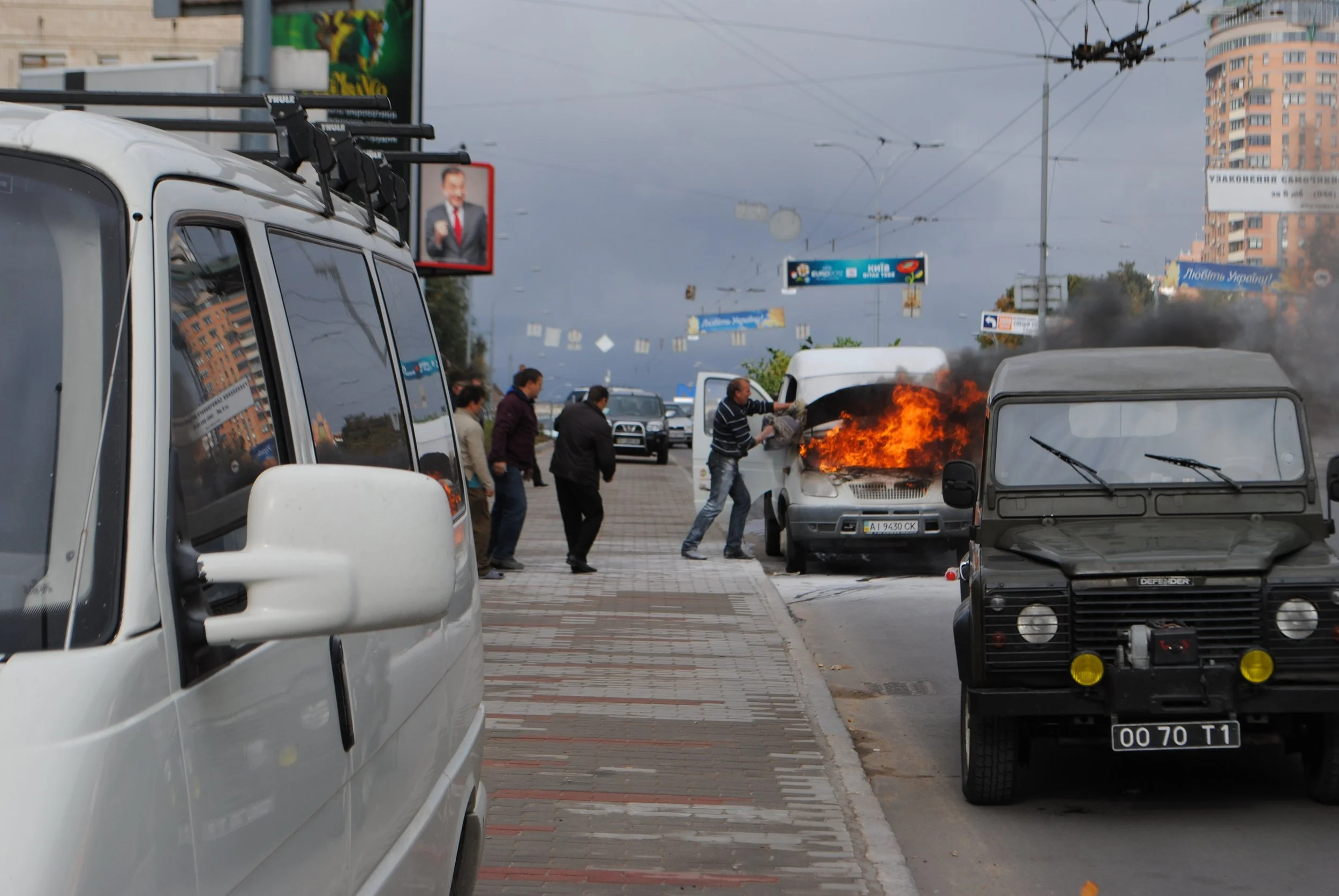 A white vehicle is parked on the side of the street with its window reflecting nearby buildings. In the middle distance, cars are burning on the street with visible flames and smoke. Several people are gathered around, some appearing to attempt to pu