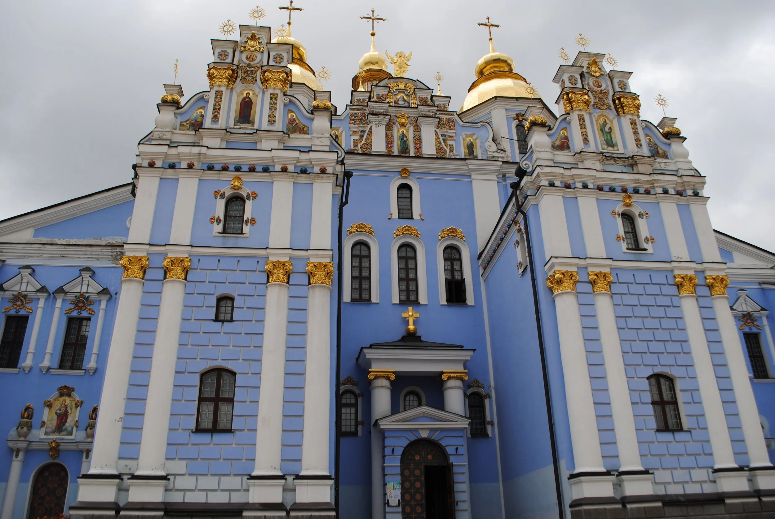 View of a blue and white cathedral with golden domes and crosses on top, adorned with religious icons and decorative details.