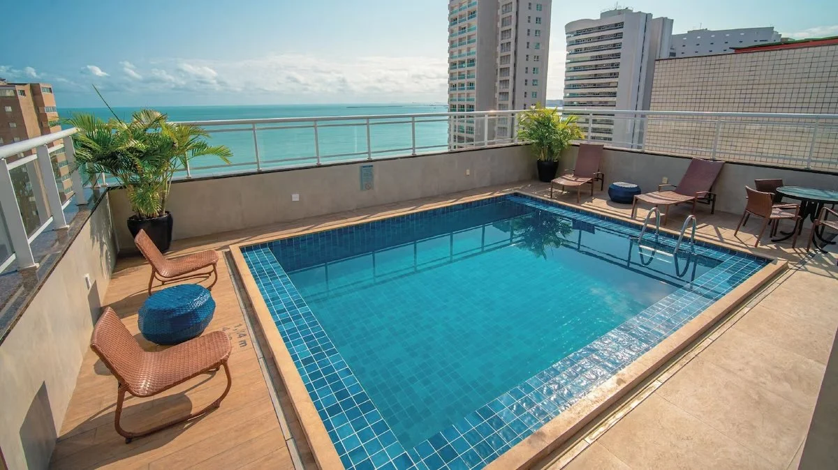 Rooftop swimming pool with city and ocean view, surrounded by lounge chairs, potted plants, and outdoor furniture.