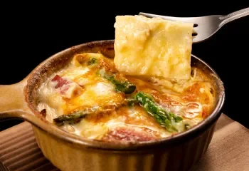 A spoon lifting a piece of cheesy baked casserole from a brown baking dish, with melted cheese, vegetables, and bacon inside.