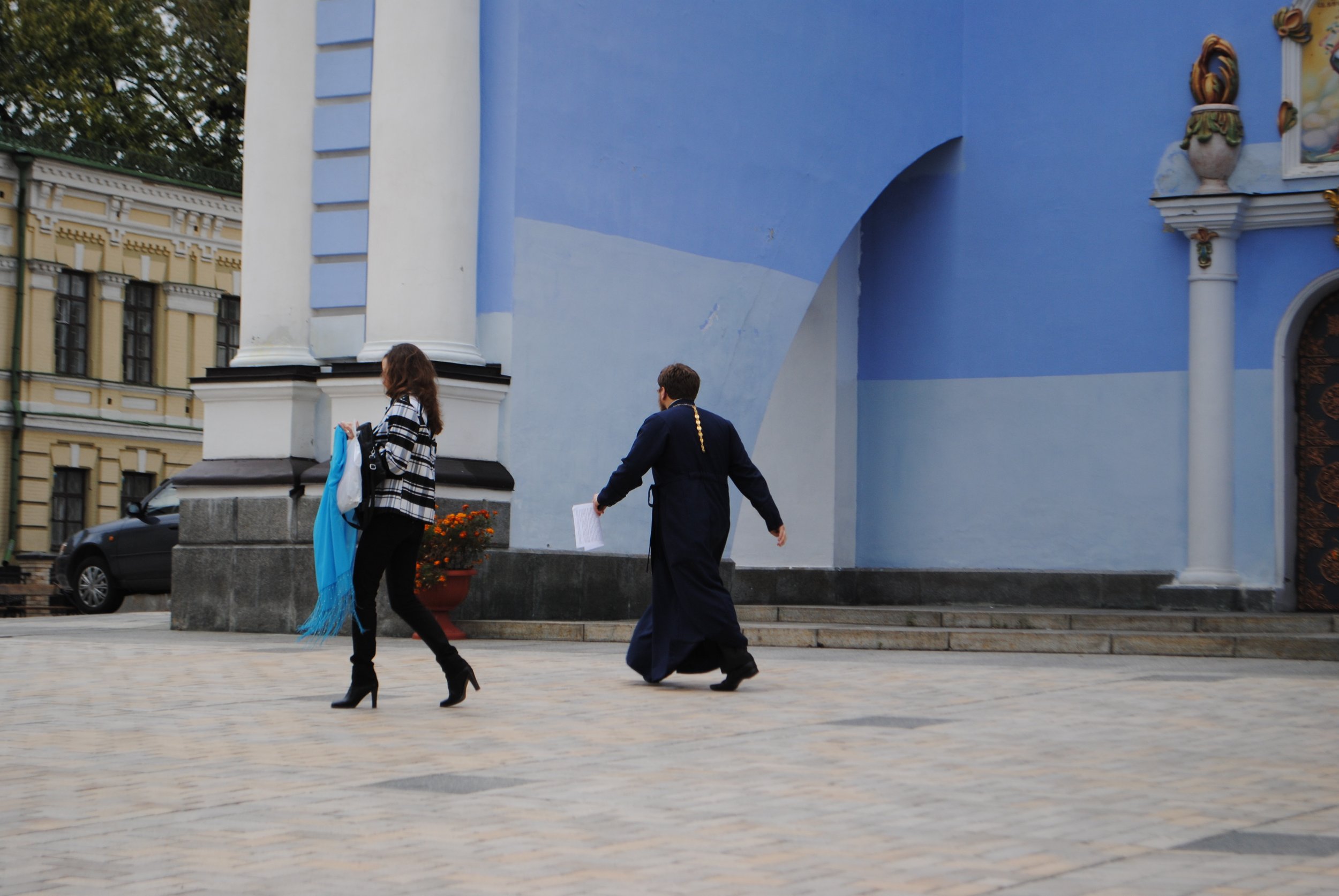 Two women and a man walking past a light blue building with ornate decorations; one woman is dressed in black pants and a checkered shirt, the other is in a formal suit, and the man is also in a suit carrying papers.