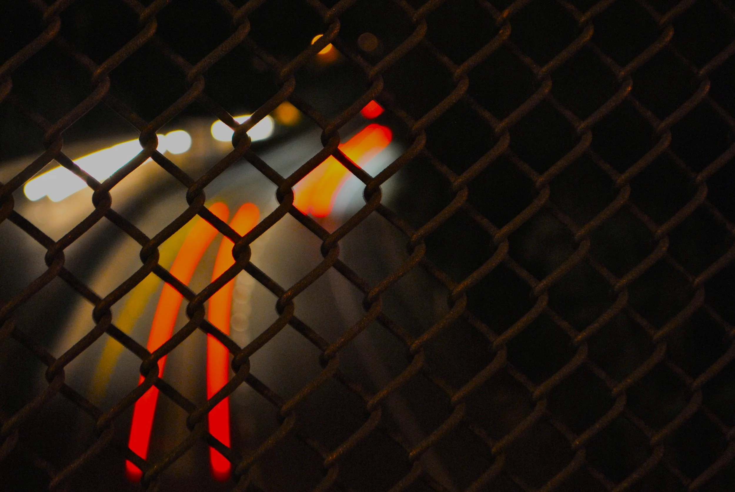 A dark image featuring a rusted metal chain-link fence in the foreground with blurred city lights in the background at night.