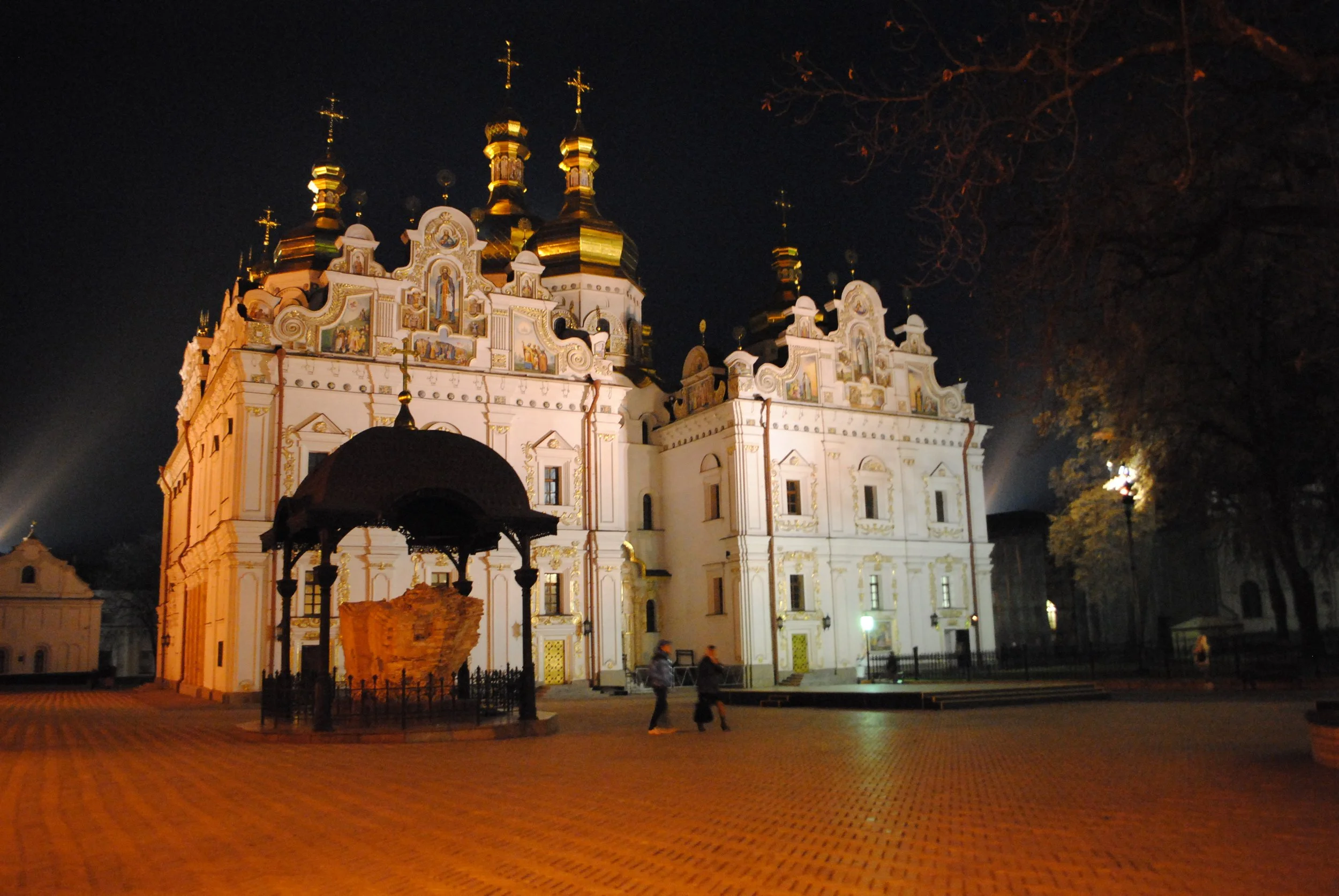 The illuminated Saint Michael's Golden-Domed Monastery at night, featuring its golden domes and intricate architecture, with a few people walking in the courtyard.