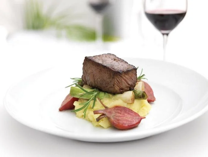 A plated serving of gourmet beef steak atop mashed potatoes, garnished with herbs and vegetables, with a glass of red wine in the background.