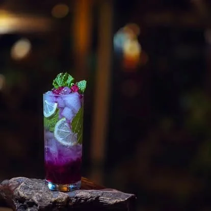 Colorful cocktail in a tall glass garnished with mint and berries on a wooden surface, blurred background.
