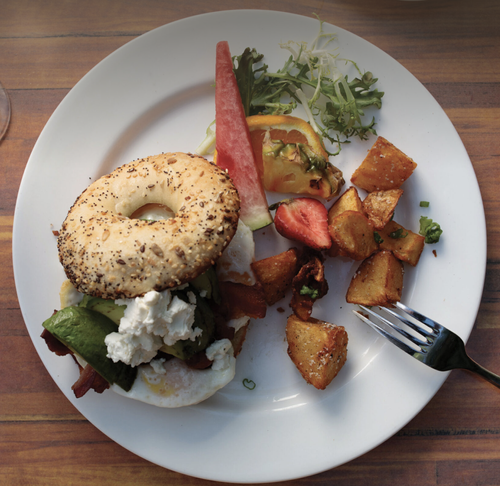 Plate with bagel sandwich topped with egg, avocado, cheese, and bacon, along with roasted potatoes, watermelon, strawberries, pineapple, and mixed greens on a wooden table.