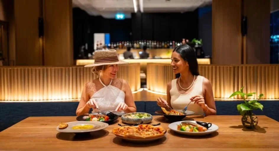 Two women sitting at a restaurant table enjoying a meal together, with various dishes including salad, pizza, and eggs, and a potted plant on the table.