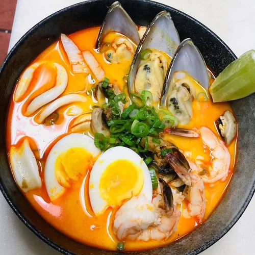 A bowl of spicy seafood soup with boiled eggs, shrimp, mussels, squid, and green onions in an orange broth, garnished with a lime wedge.