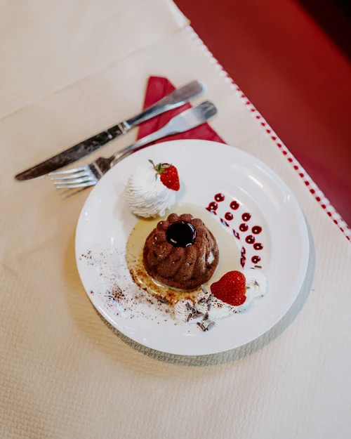 Chocolate bundt cake with cherry topping, whipped cream garnished with strawberries, chocolate shavings, and berry sauce on a white plate, with silverware and a red napkin on a beige tablecloth.