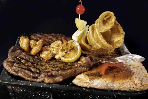 Grilled steak with garlic, lemon, cherry tomato, grilled chicken breast, fried onion rings, and potato chips on a black plate against a black background.