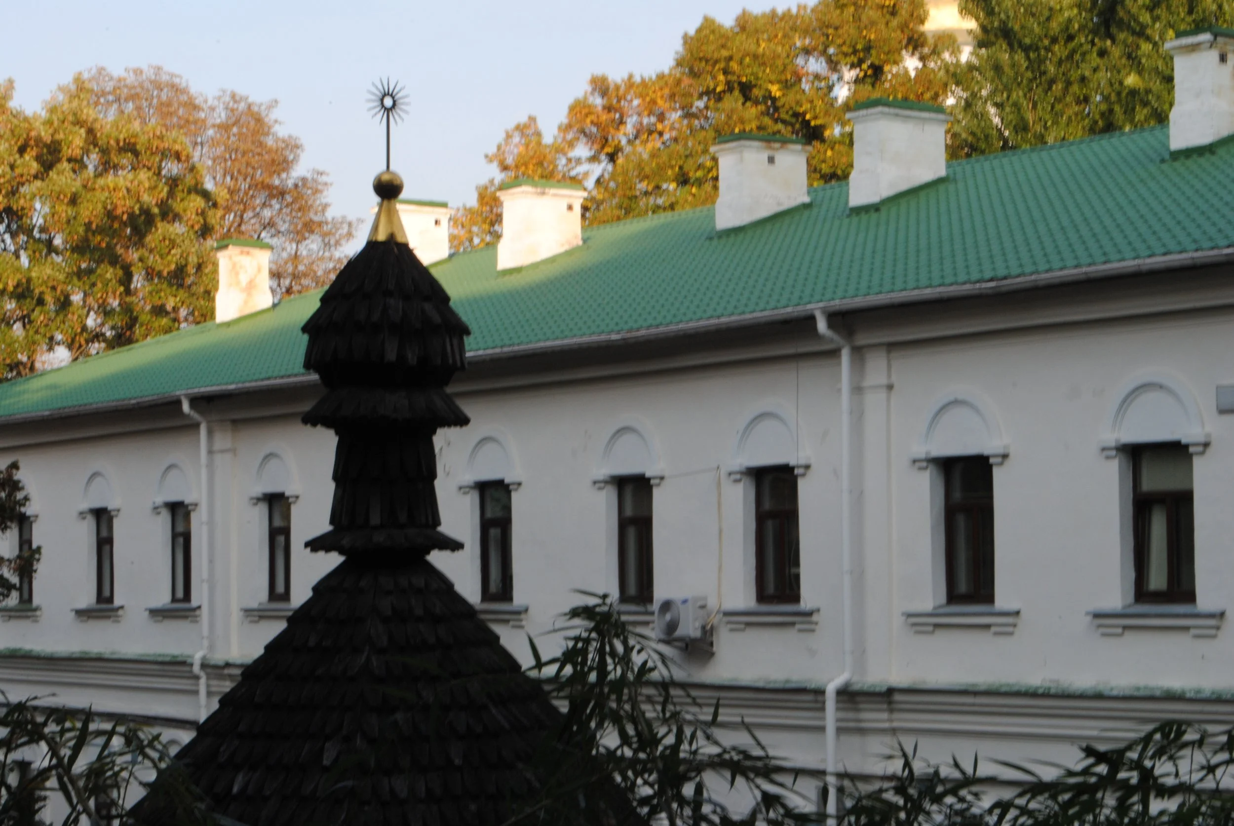 A building with white walls, green roof, and multiple chimneys, with trees exhibiting fall foliage in the background.