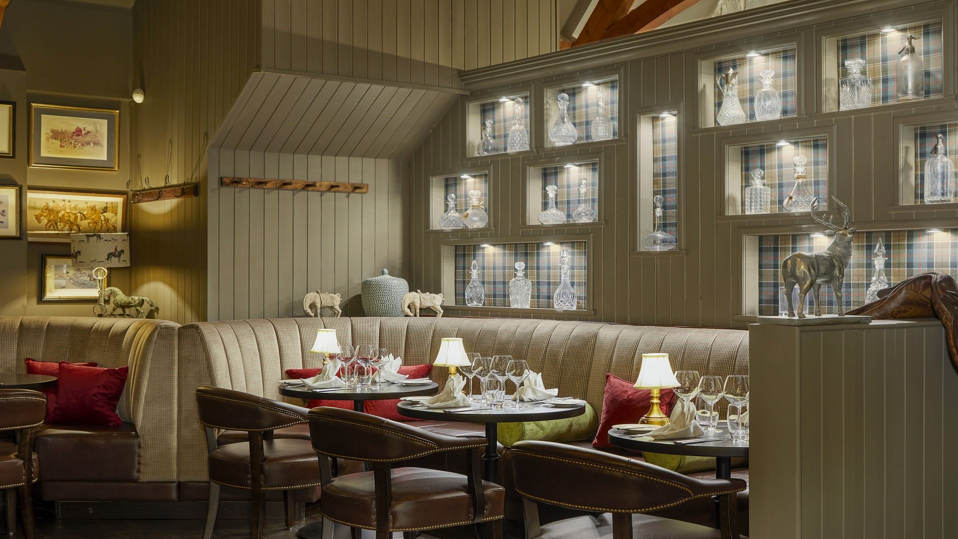 A cozy restaurant interior with a plaid wall, glass decanters on display shelves, a beige bench with red and green throw pillows, round tables set with wine glasses, napkins, and gold lamps, framed artwork on the wall, and decorative animal sculpture