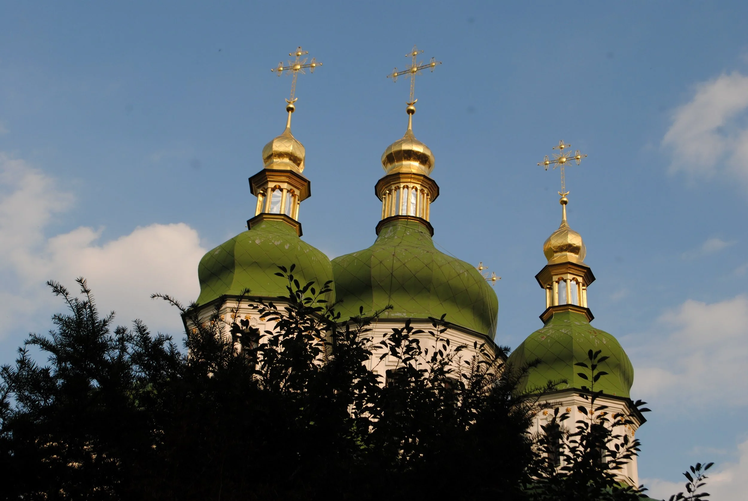 Church with green domes and gold crosses on top, partially obscured by trees in the foreground, under a partly cloudy sky.