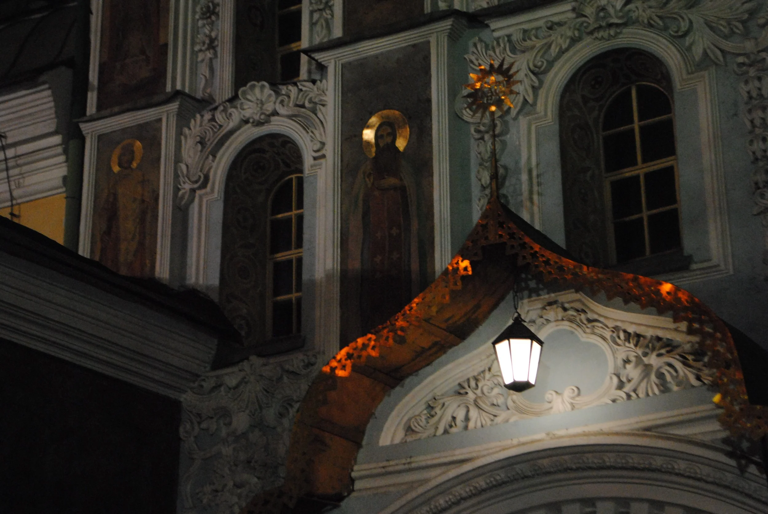 Nighttime view of an ornate church facade with detailed architectural carvings, religious statues, and a lit lamp post.
