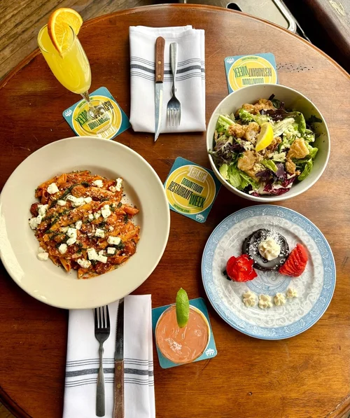 A round wooden table with a salad in a white bowl, pasta with tomato sauce and cheese, a dessert plate with a chocolate dessert, strawberries, and whipped cream, and two colorful drinks with garnishes. Silverware and napkins are arranged alongside restaurant coupons.