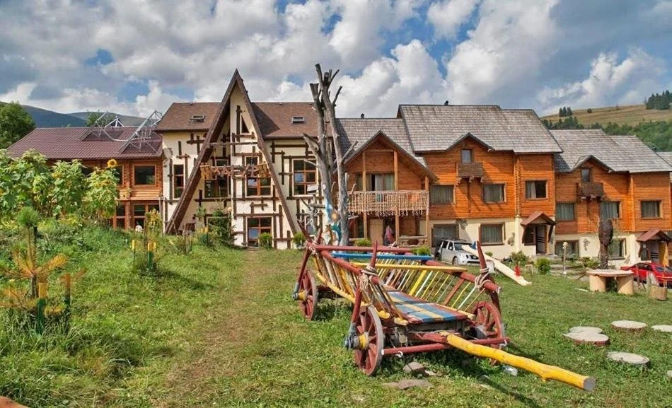 A rustic mountain village with wooden houses, a grassy yard, and a colorful, child-sized wooden cart in the foreground, with hills and cloudy sky in the background.