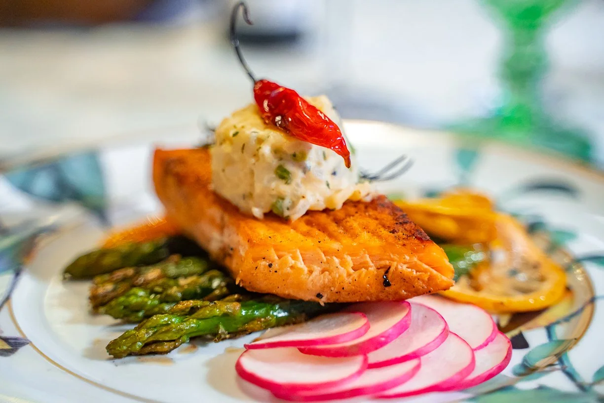 Grilled salmon fillet topped with a scoop of herbed butter and a cherry pepper, served with asparagus, radish slices, and roasted vegetables on a decorative plate.
